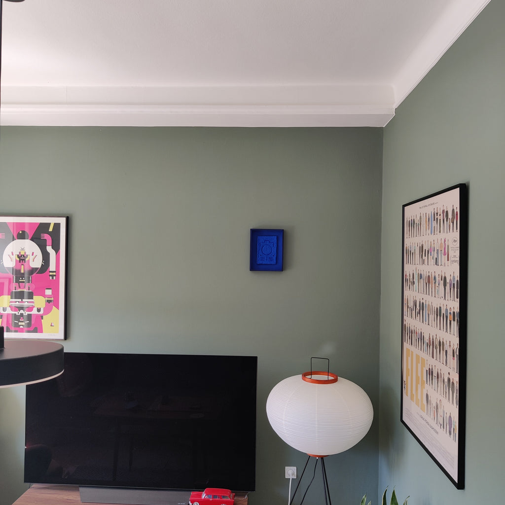 A styled interior corner of a room with sage green walls, featuring a black flat-screen TV, a white spherical table lamp with orange accents, framed wall art, and a small blue square wall hanging.
