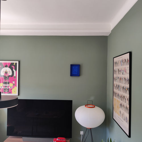 A styled interior corner of a room with sage green walls, featuring a black flat-screen TV, a white spherical table lamp with orange accents, framed wall art, and a small blue square wall hanging.