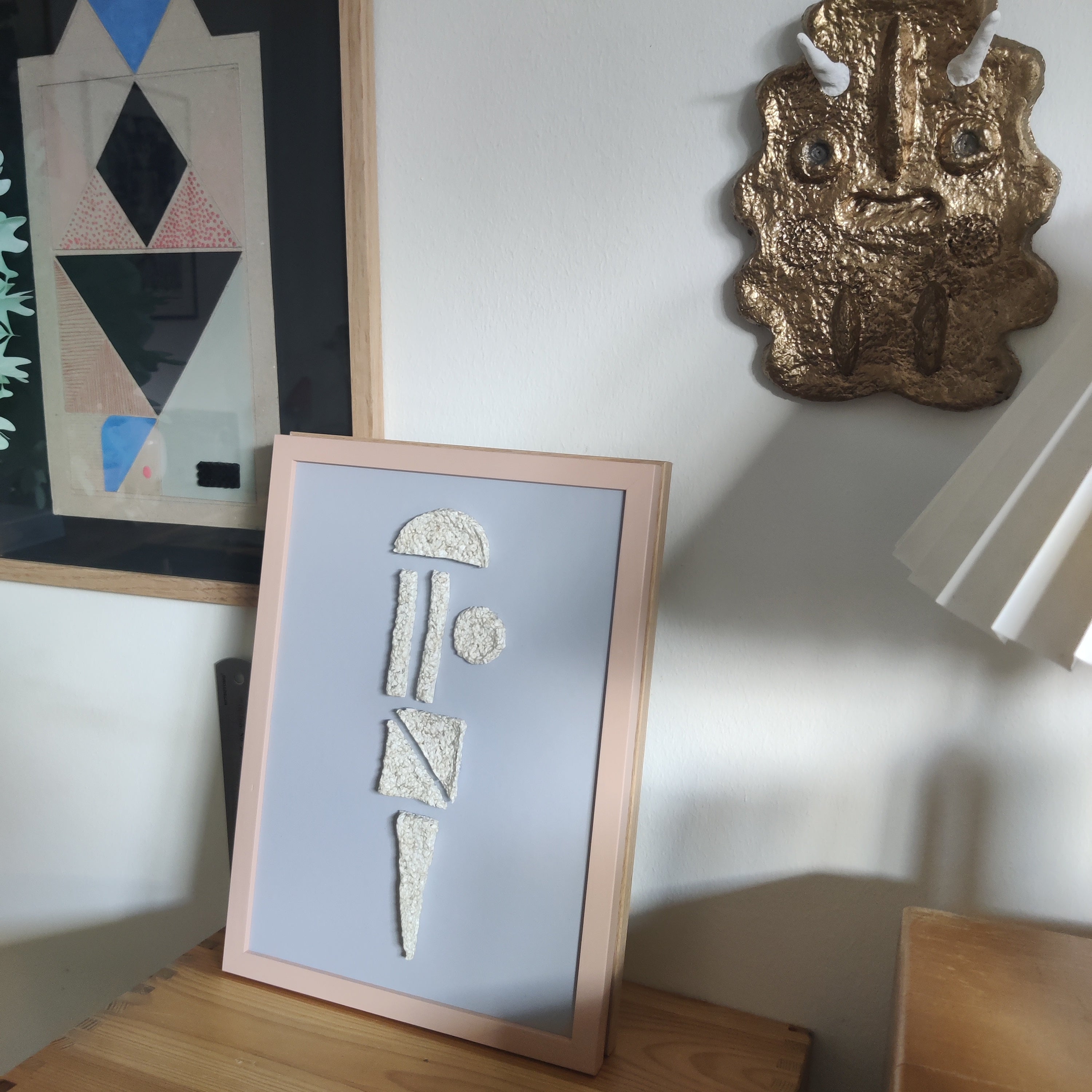 Framed artwork with abstract design on a wooden surface, with another piece of art and a lamp in the background.