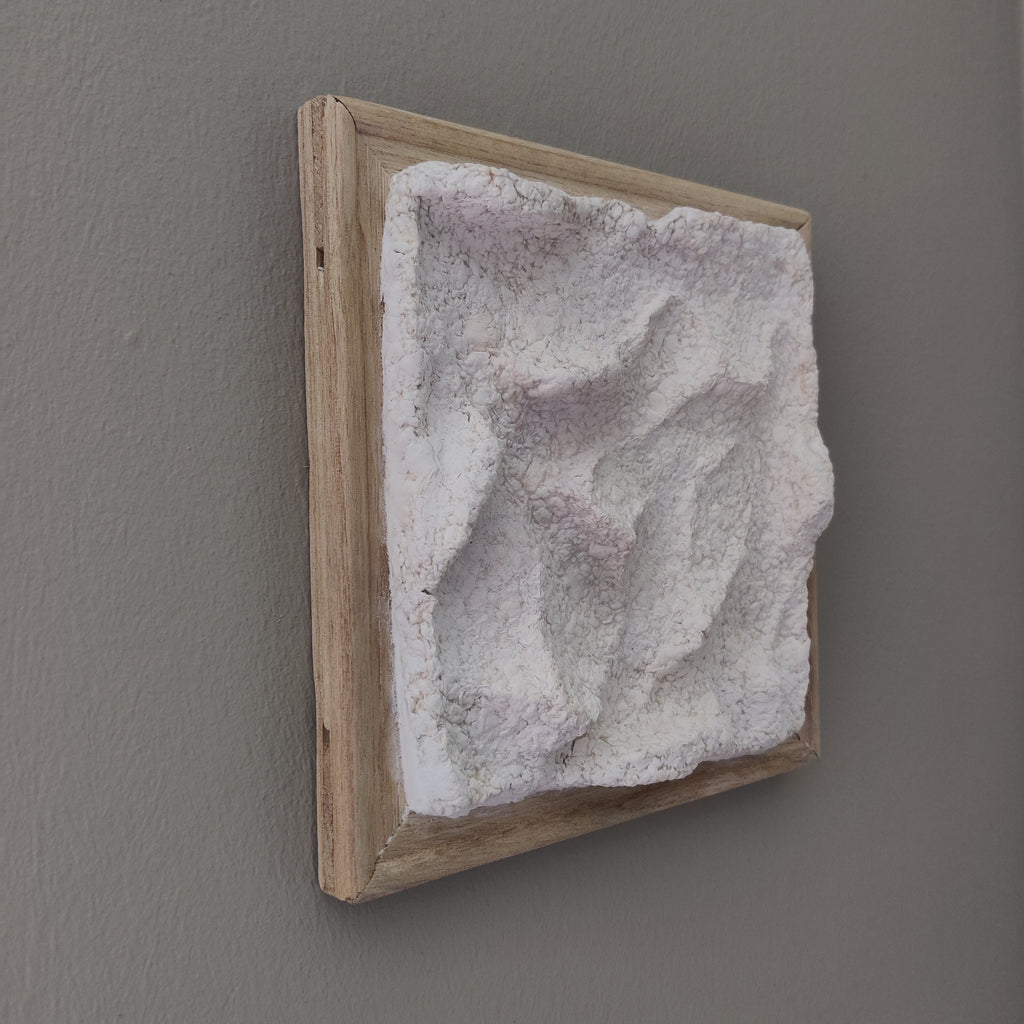 A square framed wall art piece featuring a textured, sculptural white and pale lavender surface set within a light natural wood frame, mounted on a plain gray wall.