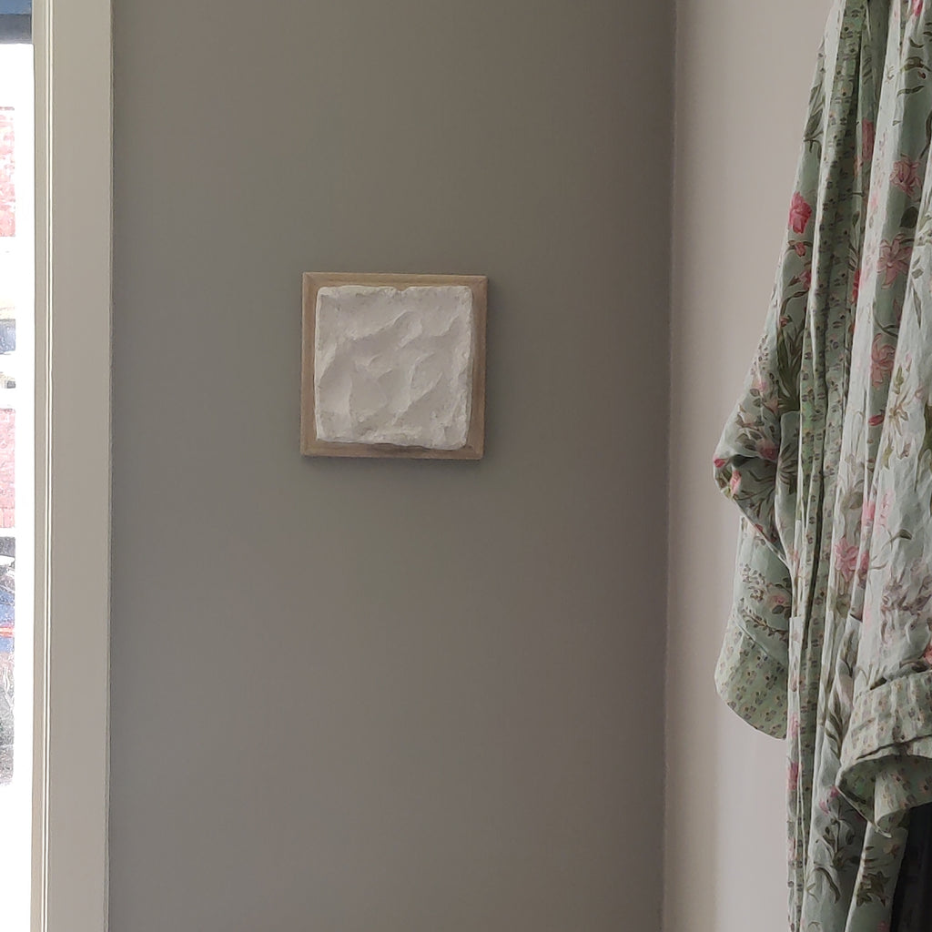 A small square framed wall art piece with a textured white center and light wood frame, mounted on a gray wall in a real interior setting with part of a floral robe visible on the right.