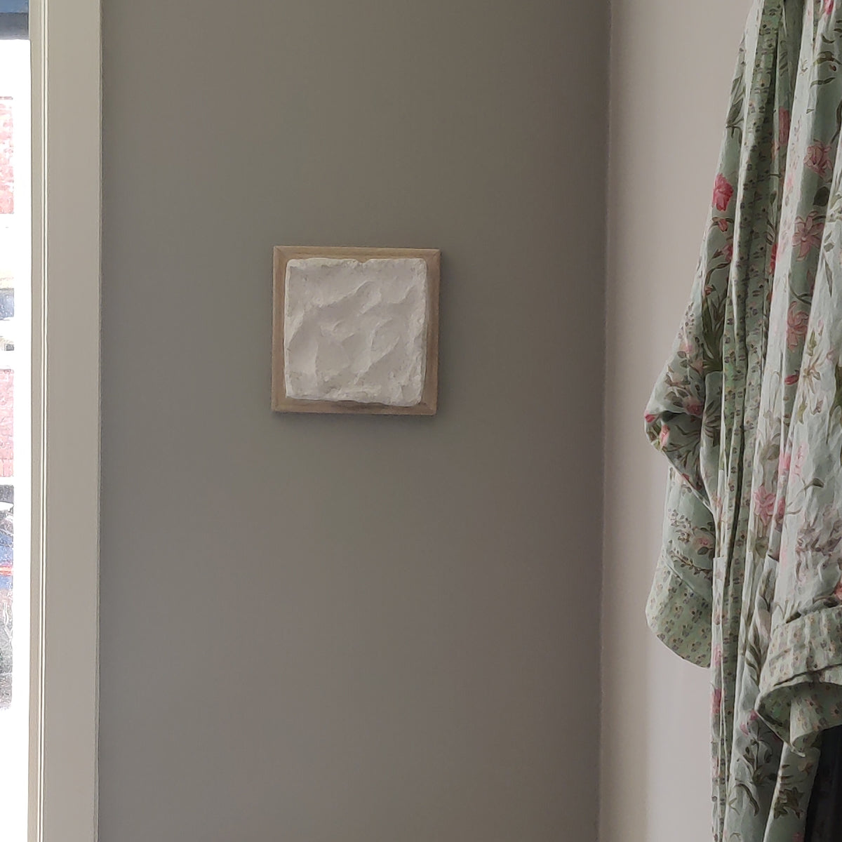 A small square framed wall art piece with a textured white center and light wood frame, mounted on a gray wall in a real interior setting with part of a floral robe visible on the right.