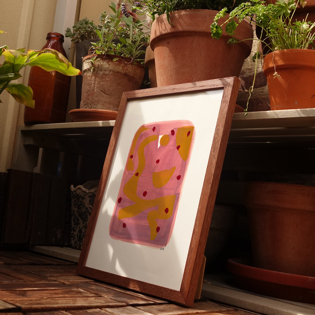 A framed abstract art print with pink and yellow shapes, leaning on a wooden surface in a styled indoor setting with potted plants in the background.